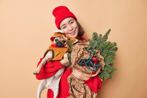 Free photo lovely pleased brunette woman poses with pug dog and holiday attributes prepares for christmas or new year celebrations wears red sweater and hat enjoys festive time with favorite pet poses indoor