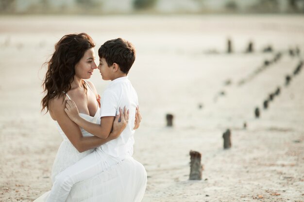 Lovely portrait of mother and son on the sand, dressed in the white clothes, looking each other in the eyes