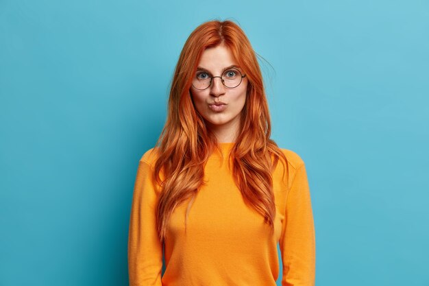 Lovely redhead young woman in round glasses keeps lips folded wants to kiss someone dressed in orange jumper looks directly.