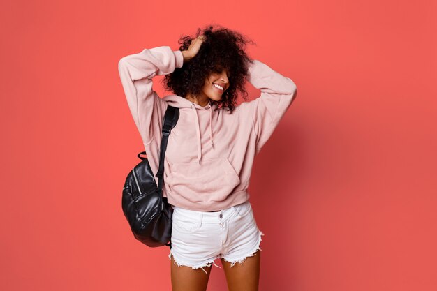 lovely  sexy black woman in stylish hoodie   with back pack posing on pink background and playing with curly hairs.