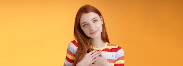 Free photo lovely silly cute redhead young european girl tilting head look gently smiling tender camera press palms heart thankful receive nice gift gazing gratitude happiness standing orange background