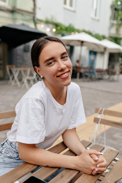 Lovely smiling lady with dark collected hair wearing white tshirt is posing at camera with wonderful smile while resting outdoor in cafeteria