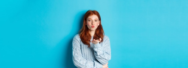 Free photo lovely teen girl in sweater blowing air kiss pucker lips and staring at camera standing against blue