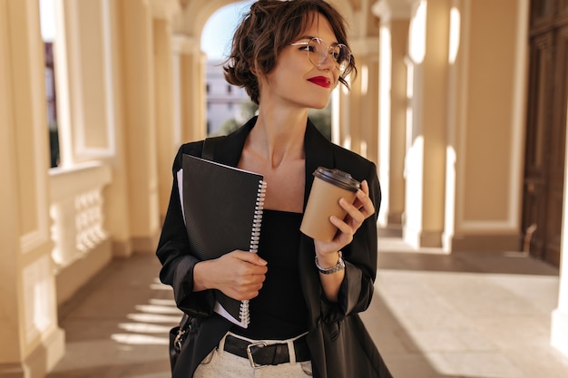 Free photo lovely woman in glasses holding cup of tea and notebook outside. brunette woman with red lips in black jacket smiling outdoors.