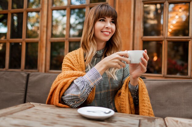 Lovely woman have a coffee break in cozy cafe with wood interior, talking by mobile phone. Holding cup of hot cappuccino. Winter season. Wearing elegant dress and yellow plaid.