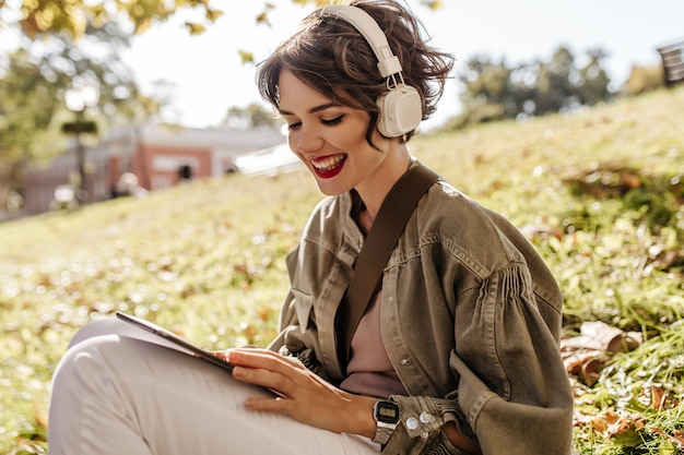 Free photo lovely woman in jacket and headphones sitting on grass outdoors. happy woman with curly hairstyle holding smartphone outside.