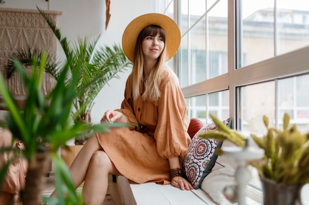 Lovely woman in linen dress and straw hat posing in boho style apartment