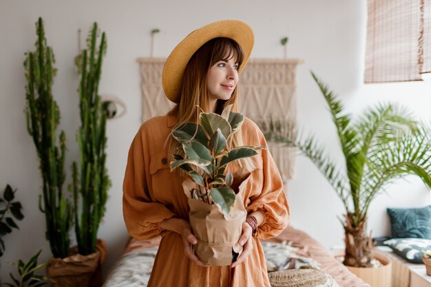 Lovely woman in linen dress and straw hat posing in boho style apartment