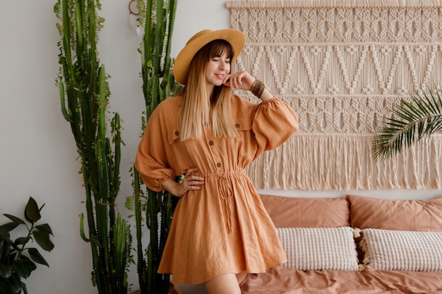 Free photo lovely woman in linen dress and straw hat posing in boho style apartment