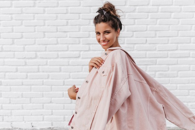 Free photo lovely woman in pink denim jacket smiling and posing on brick light wall