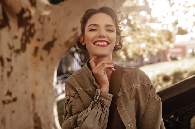 Lovely woman with brunette short hairstyle in denim jacket smiling outdoors. Charming woman with red lips posing outside.