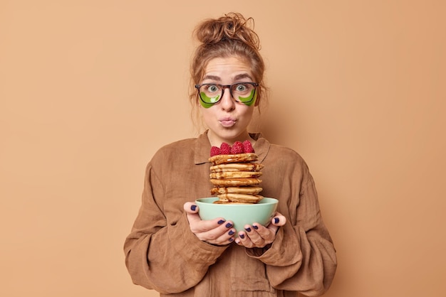 Free photo lovely wondered woman keeps lips folded applies green hydrogel patches under eyes to reduce puffiness dressed in nightwear holds delicious pancakes with syrup has tasty breakfast applies beauty pads