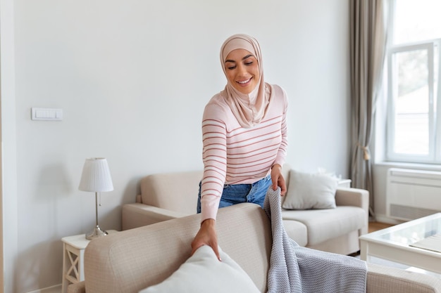 Lovely young Arabic Muslim woman putting soft pillows and plaid on comfy sofa making her home cozy and warm