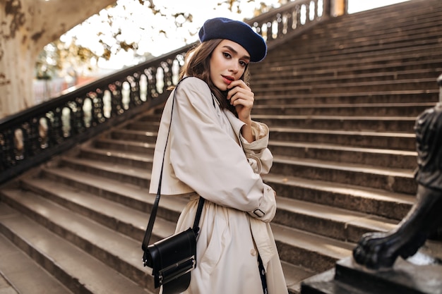 Free photo lovely young parisian woman with brunette hair in stylish beret, beige trench coat and black bag, standing on old stairs and sensitively posing outdoors