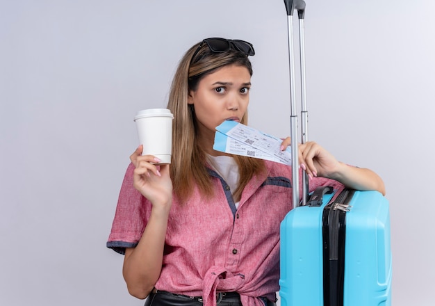 A lovely young woman wearing red shirt and sunglasses looking while holding plane tickets with blue suitcase on a white wall