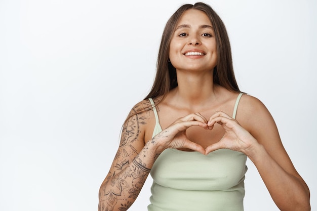 Free photo lovely young woman with tattoos on arm, showing heart sign and smiling caring at camera, white background