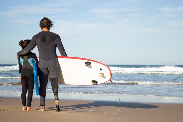 Loving father with mechanical leg with daughter on beach.  Back view of mid adult man and little dark-haired girl carrying surfboards, looking at water. Family, leisure, active lifestyle concept