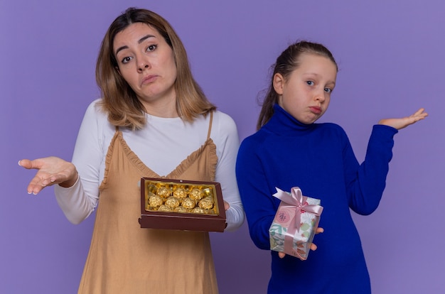 Loving mother with box of chocolates and daughter holding present looking confused raising arms celebrating international women's day standing over purple wall