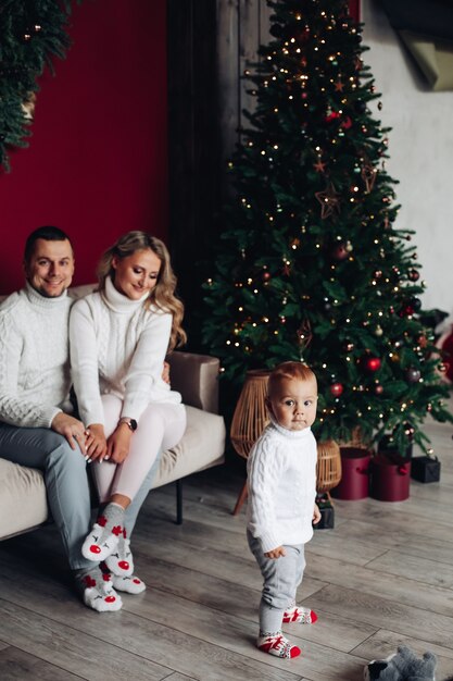 Loving parents in white clothes sitting on couch near Christmas tree while watching their kid