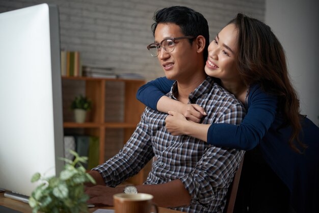 Loving wife hugging her husband from the back while he is working at computer