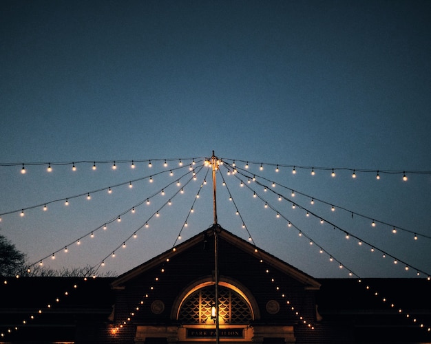 Low angle shot of strings of light bulbs attached to a pole in front  of a park pavilion at dusk