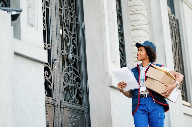 Free photo low angle view of smiling african american postal worker carrying packages while making delivery in residential district