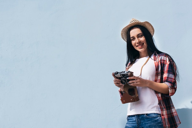 Low angle view of smiling woman wearing hat holding retro camera standing against sky