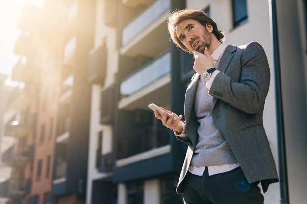 Low angle view of young businessman thinking while text messaging on mobile phone in the city