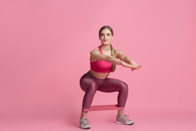 Lower body. Beautiful young female athlete practicing , monochrome pink portrait. Sportive fit caucasian model with elastics. Body building, healthy lifestyle, beauty and action concept.