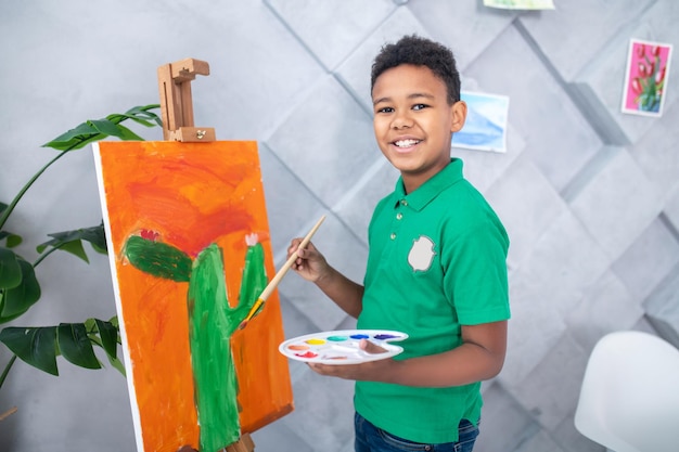 Free photo lucky day. dark-skinned cute school-age boy in green tshirt with palette and brush in his hands near easel standing smiling at camera in room