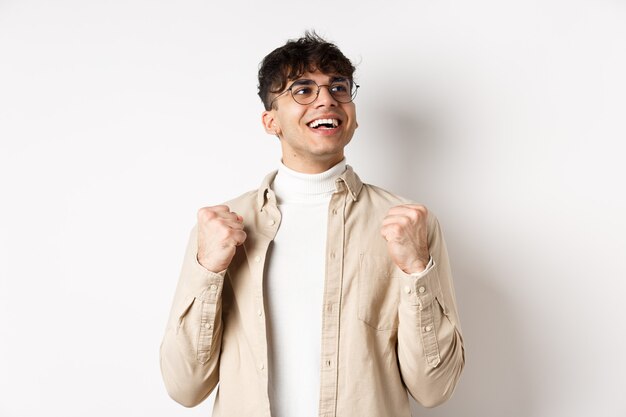 Lucky young man say yes with relieved face, smiling and making fist pump, triumphing of success, winning prize, standing on white background after achieving goal