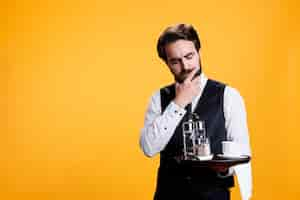 Free photo luxury butler thinking about ideas, preparing to serve customers with food or drinks in yellow studio. stylish person with waiter occupation being thoughtful with tray in hand on camera.