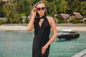 Free photo luxury sexy attractive woman dressed in black dress posing on pier in luxury resort hotel, wearing sunglasses, summer vacation, tropical beach