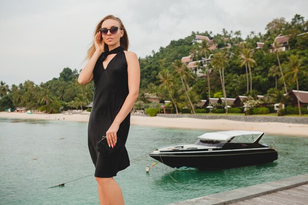 Free Photo luxury sexy attractive woman dressed in black dress posing on pier in luxury resort hotel, wearing sunglasses, summer vacation, tropical beach