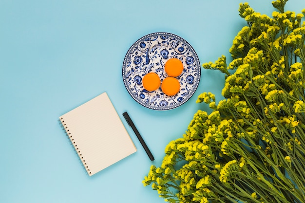 Free photo macaroons on ceramic plate; spiral notepad; pen and bunch of yellow flowers on blue background