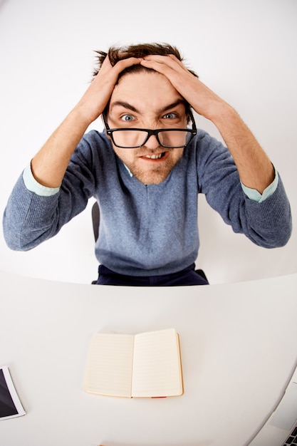 Mad and distressed young pressured man, toursle hair and grimacing angry as work, sit desk can't think-up ideas