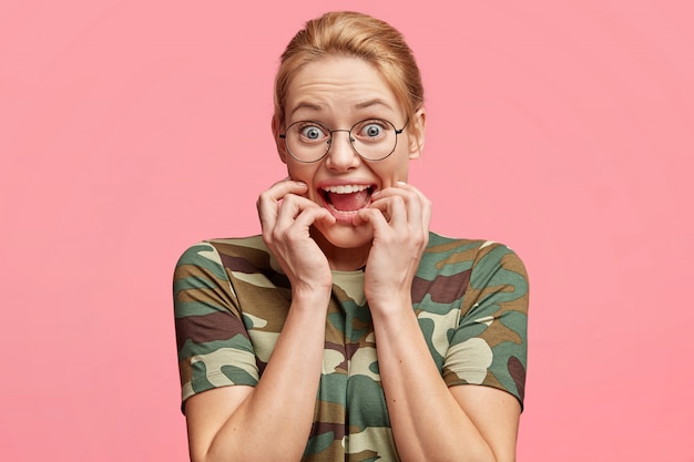 Free photo mad surprised female model with eyes popped out, stares through round spectacles, bites finger nails, feels nervous and excited, reacts on something very actively, isolated over pink