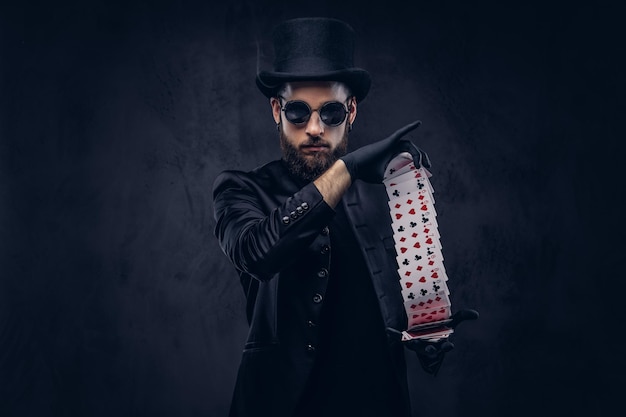 Free photo magician in a black suit, sunglasses and top hat, showing trick with playing cards on a dark background.