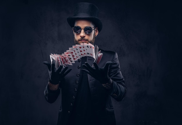 Free photo magician in a black suit, sunglasses and top hat, showing trick with playing cards on a dark background.