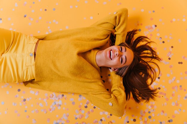 Free photo magnificent girl in yellow pants lying on purple confetti. overhead portrait of enthusiastic emotional woman.