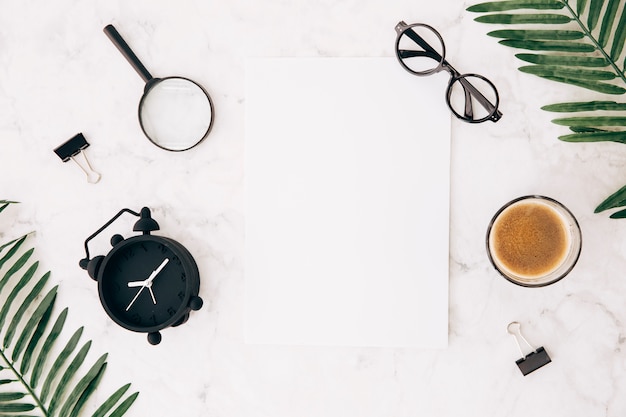 Magnifying glasses; alarm clock; eyeglasses; coffee; bulldog clip and leaves with blank white paper on marble textured backdrop