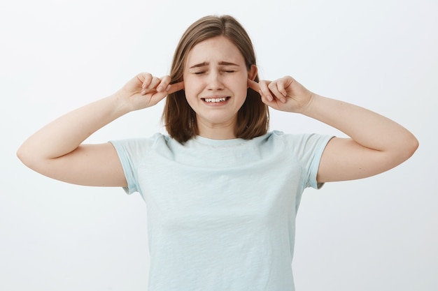 Make this sound stop. Unhappy insecure crying cute girl with brown hair closing eyes and clenching teeth making displeased sad expression covering ears with index fingers not hear parents fighting