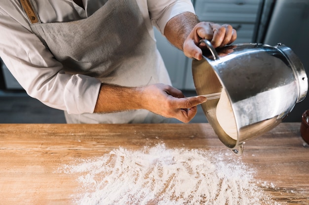 Free photo male baker pouring knead dough from container on wooden table dusted with flour