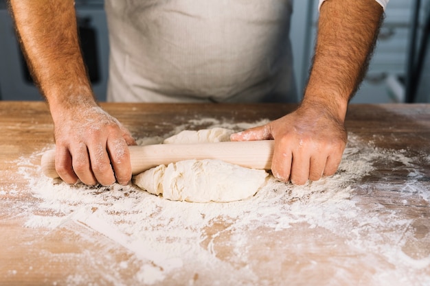 Free photo male baker's hand flattening dough with rolling pin on wooden table