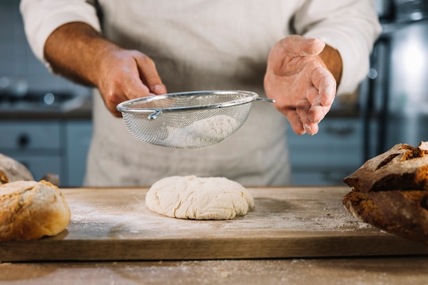 Free Photo male baker sifting the wheat flour through steel sieve over knead dough