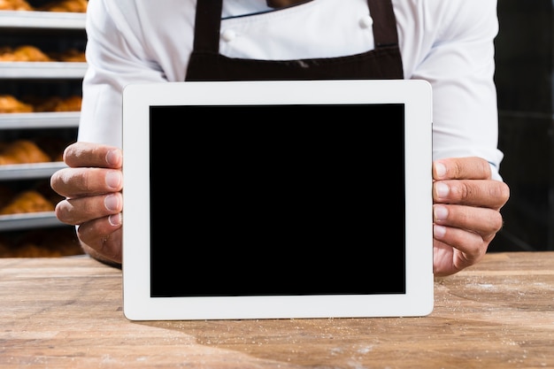 A male baker in uniform holding small blank digital tablet on wooden table