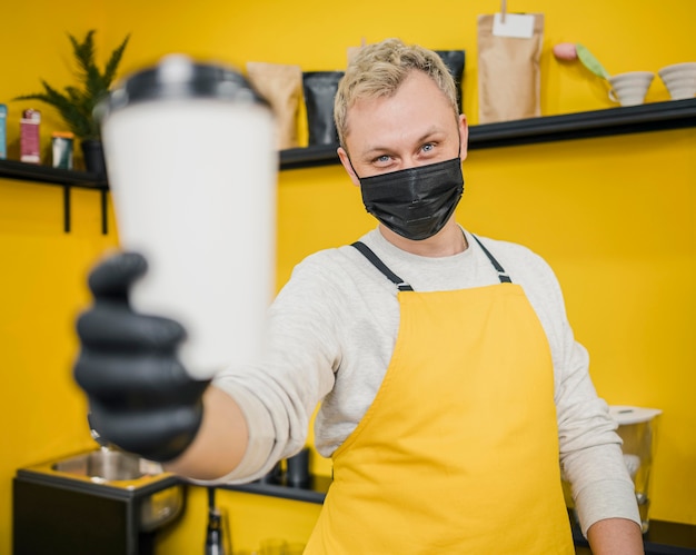 Male barista with medical mask holding coffee cup