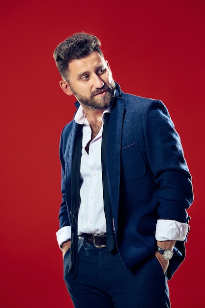 Male beauty concept. Portrait of a fashionable young man with stylish haircut wearing trendy suit posing over red background.