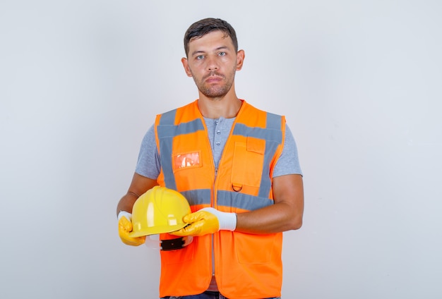 Male builder holding helmet in his hands in uniform, jeans, gloves, front view.