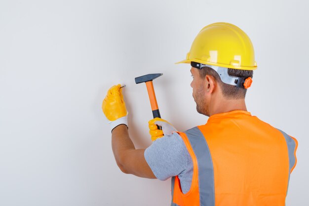 Male builder in uniform, helmet, gloves hammering nail into wall , back view.